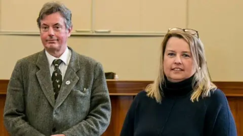 Sarah Caldecott Councillors Richard Bell and Amanda Hopgood standing next to each other and looking directly at the camera.
Bell has short grey hair and wears a grey suit jacket with white shirt and green tie.
Hopgood has medium length straight blond hair and her glasses are on the top of her head. She wears a navy blue jumper. 
