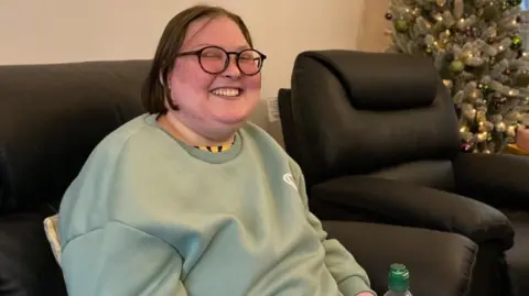 A young woman with glasses smiles as she sits infront of a Christmas tree.