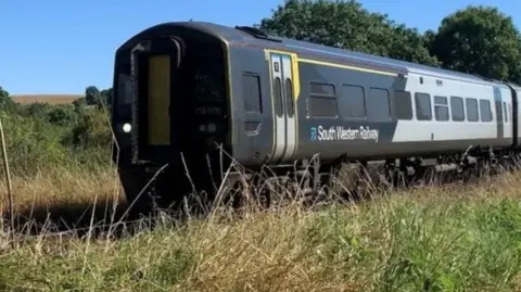 A South Western Railway train passes parched vegetation.