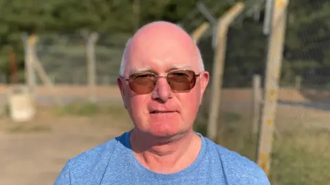Luke Deal/BBC Mick Coppin smiles at the camera. He is largely bald with some grey hair around the sides of his head. He wears sunglasses and a blue T-shirt.