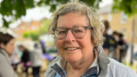 Tom Jackson/BBC Anne Miller has short blonde curly hair. She is wearing light grey framed glasses, a blue and white striped jumper and a white jacket. Behind Miller is blurred, but there are several people congregating on a field.