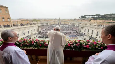 Getty Images The Pope waves at people on St Peter's Square in Vatican City. He camera is behind him as he stand son a balcony. The people are below.