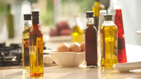 A collection of bottles of cooking oils and vinegars on a kitchen counter. There is a bowl of eggs in the centre and a gas hob in the left background.