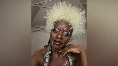 A person in bold avant‑garde makeup with bright blue eyes, a large white beaded wig, and metallic costume accessories, standing in a tent-like backstage setting