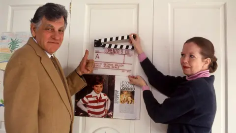 Getty Images A middle-aged couple stand in front of a set of white doors holding a movie clapper board and a magazine cutting. On the left,a man with a mop of dark gry hair wearing a fawn blazer and given the thumbs up. On the right a woman with brown hair, tied back, wearing a blue and pink sweater. The clapper board is white with the words "Lorenzo's Oil, Director George Miller, Camera John Seale" printed on it in red letters. The magazine clipping includes a picture of a young boy wearing a red and white striped rugby shirt. 