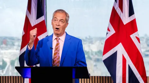 Nigel Farage in a blue suit and pink shirt and tie, standing in front of a blue background with a union flag in the background