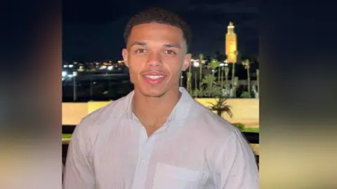Sam Rushton Young man with short hair wears white shirt and stands posing for a photo in the evening with palm trees in the background