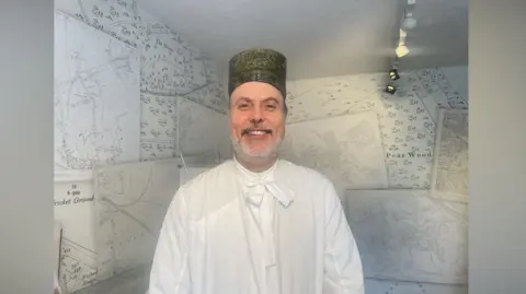 A man smiles as he poses for a photo. He is wearing a white garment and a green circular brimless hat.