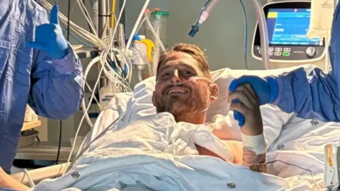 A man in a hospital bed with a breathing tube, medical equipment can be seen in the background