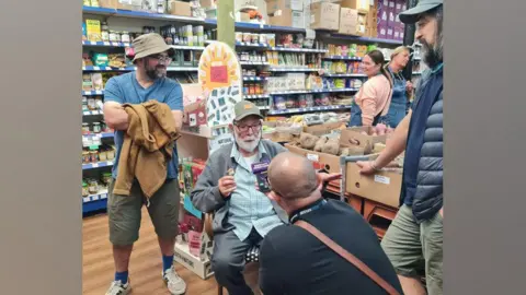 Matta's International Foods Mr Matta is sat on a chair in his shop as he is interviewed by BBC Radio Merseyside's Kev Duala. His two sons Deepak and Dalip stand on either side of him.
