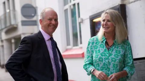 Jersey Overseas Aid Vice Admiral Jerry Kyd CBE pictured smiling with Jersey’s Minister for International Development, Deputy Carolyn Labey. Deputy Labey is wearing a green and white shirt, she has blonde hair. Vice Admiral Kyd is wearing a pink suit shirt, a navy blue patterned tie and a black blazer on top.
