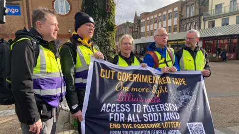 The group of campaigners are pictured together as they set off on their walk. Five people are pictured and they are all wearing high-viz vests. They are holding a banner that reads: "Cut government red tape. One more light. Lillia's walk. Access to tofersen for all SOD1 MND. End the postcode lottery. From Southampton General Hospital."