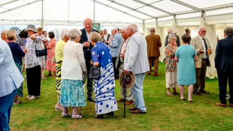 A wide shot of a garden party. There are hundreds of people gathered inside a large marquee.