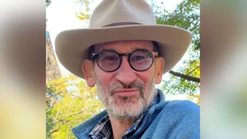 Jimmy Walker A man with a grey and black beard, glasses, blue jumper and a large brown felt hat looking straight down the camera lens with trees and sky in the background.