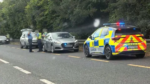 Garda officers and garda cars surround a small car on the roadside of the N2  near Carrickmacross. The officers are all wearing blue uniforms, but two are wearing yellow high-viz vests over their shirts.  The boot of a hatchback vehicle behind them is open. 
