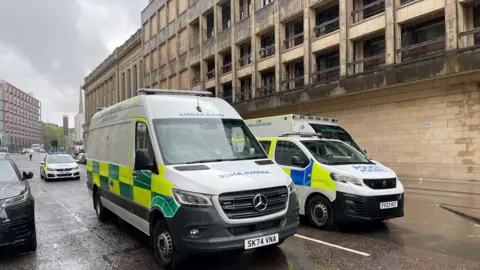A police van, car and ambulance sitting outside the Mitchell library building, with a police officer walking around the scene in the distance. 