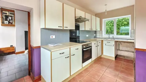 Paul Fosh Auctions A kitchen with white kitchen units and purple walls. There is also brown/ orange tiles on the floor and blue and white tiles on the walls. 