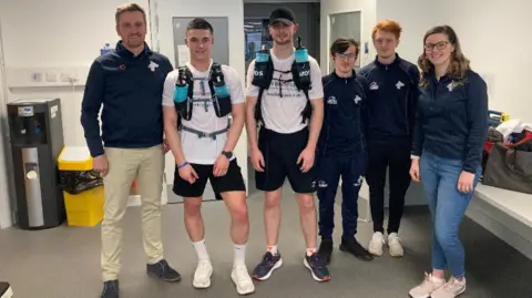 James and Greg are wearing T-shirts and shorts with kit on their back. They are standing with students and a lecturer at the Walsall campus.