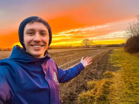 Jack Silkstone A man wearing a purple cat with Universal theme park branding is stood on a large field at sunset