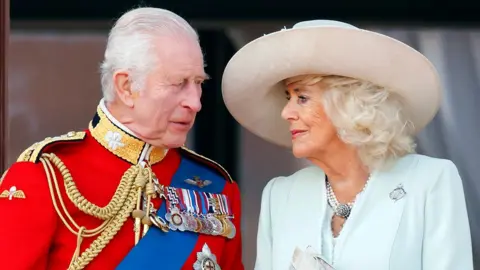 Getty Images O rei Charles, à esquerda, vestindo um uniforme vermelho com medalhas presas, encara a rainha Camilla, vestindo uma jaqueta azul-clara e um chapéu bege. Ela também está olhando para ele