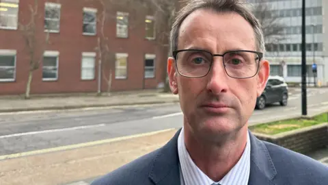 Ben Parker/BBC Lee Nunn wearing a blue suit, white shirt and blue tie. He has glasses on with brown hair. In the background is Ipswich Magistrates' Court