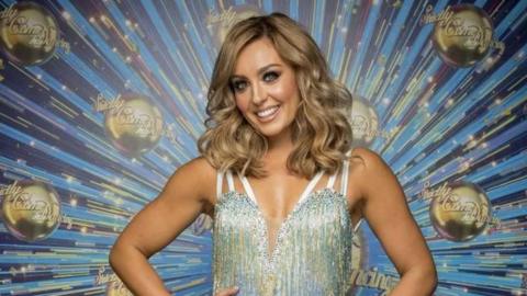 Amy Dowden looking at the camera in a silver sparkly dress with Strictly Come Dancing logo behind her