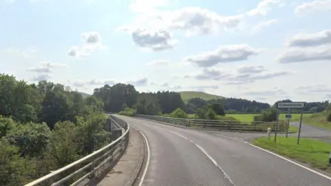 Google A road with barriers on either side. There are also trees in full leaf on both sides of the road, and a hill in the distance. There is a road sign on the right pointing to Commonside.