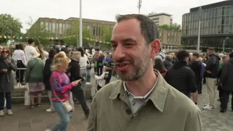 Jonathan Harper speaking outside in Bradford. He is wearing an overshirt and has a short beard. There are lots of people gathered in the background.