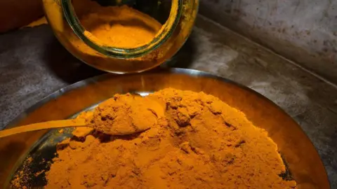NurPhoto via Getty Images Light yellow turmeric powder spread across a large metal tray, with a steel spoon partially buried in the spice. An open glass jar tipped on its side spills more turmeric into the tray.