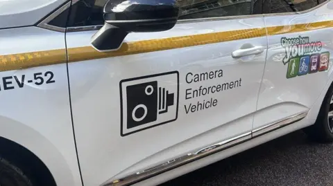 Branding and markings on the side of a white car, including a speed camera sign and the words Camera Enforcement Vehicle