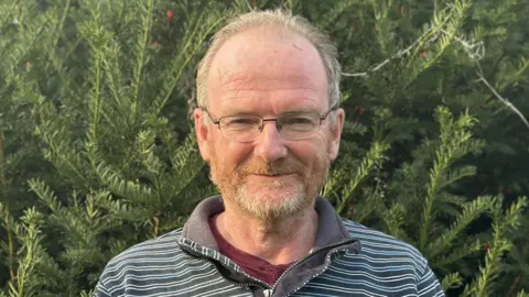 Philip Parker has short grey hair and is wearing black framed glasses, a burgundy T-shirt and a blue jumper. He is standing in front of a large tree.