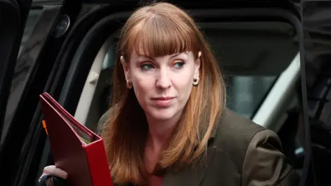 Angela Rayner, a woman with short red hair and a fringe wearing a brown jacket, steps out of a black car holding a red folder.