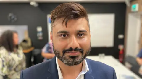 A man looking at the camera and smiling. He has light brown hair with a quiff. He has a beard with both black and grey hair. He is wearing a white shirt with the top button undone and a dark blue suit jacket.