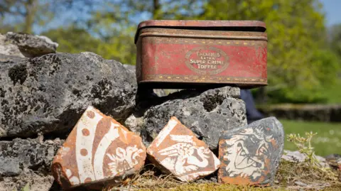English Heritage Three patterned brown tiles lying in front of a collection of stones with a battered old tin behind them with the words Thornes Toffees on it