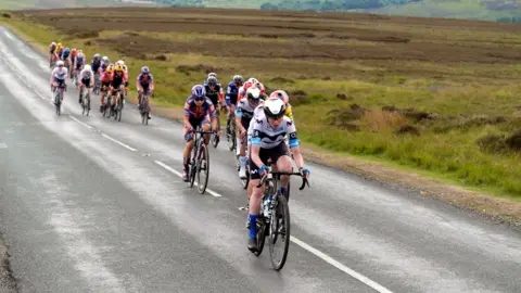 PA Media A number of cyclists, dressed in different coloured outfits, ride along a single-carriageway road in the middle of the countryside.