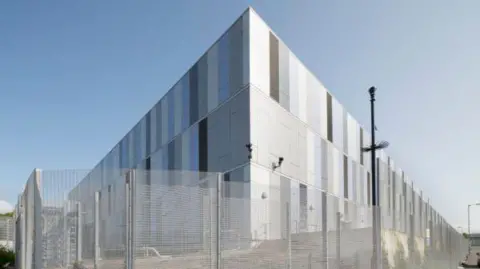 A large silver data centre building, with a mesh fence surrounding it.