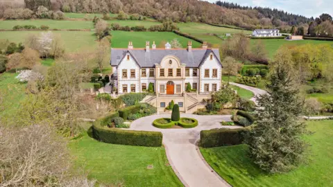 Fine and Country Exterior shot of the mansion seen with the driveway leading up the entrance. The bushes around the property are perfectly groomed.