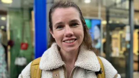 Fiona Kellagher has brown hair and is smiling. She is wearing a fleecy white jacket and the yellow straps of a rucksack are over her shoulder. 