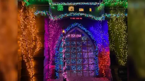 Castle fabrication lit up with bright lights, seen from the front, through the front archway with its portcullis. There are several trees in front of it which each have a different colour of Christmas lights in them. They are yellow, blue, pink and orange. A garland that runs around the edge of the castle has green lights.