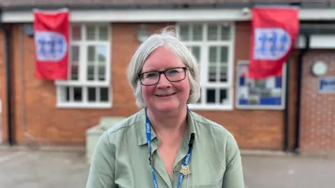 BBC News A woman with short grey hair, wearing black-rimmed glasses and a green shirt, smiling in front of a school building. At either side of her there is an England football badge flag. 
