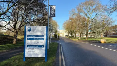 Martin Heath/BBC A roadway towards a two-storey stately home. There is a blue-framed sign on an A board to the left of the picture, with directions to the hall and other areas. There are trees on both sides of the road, and a yellow salt bin to the right.