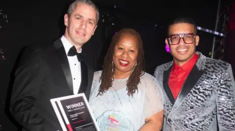 MBCC Three people stood against a black background. The man on the left is wearing a black tuxedo, a woman in the middle is wearing a blue dress and holding a glass award and certificate which says winner. A man on the right is wearing a silver suit