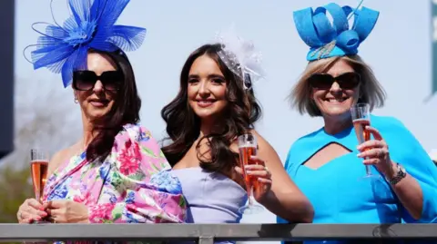 PA Media Three women stand along a railing. One is wearing a pink and blue floral dress, and blue fascinator, another is wearing a lilac dress and the third is wearing a blue dress and blue fascinator. They are each holding a glass of pink champagne.