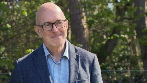 A bald man with glasses, wearing a suit jacket and shirt, smiles while standing in front of leafy green trees 