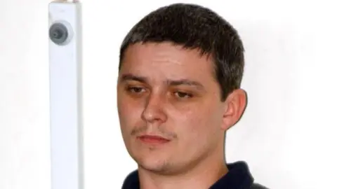 PA A file image of Ian Huntley. He has short, dark hair, a light moustache and some stubble on his narrow chin. His brown eyes look down as he stands near a brightly lit white wall.