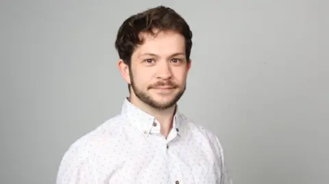 BMA chris smith wearing a light-coloured, patterned button-up shirt stands against a plain grey background in a studio-style portrait setting