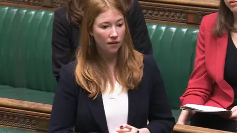 House of Commons Labour MP for Hastings and Rye, Helena Dollimore, pictured in the House of Commons chamber. She is wearing her ginger hair down, and is dressed in a navy suit jacket and white undershirt.