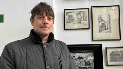 Scott Duffin is wearing a dark grey coat, with collar turned up. He is standing in front of prints which ar part of the Coventry Market exhibition