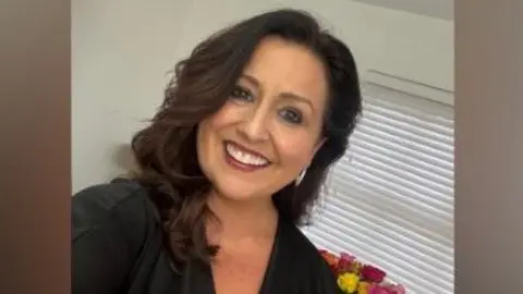 Lisa McFarland A woman with long brown hair is smiling into the camera. She is wearing a black v neck top. Behind her is a colourful bunch of flowers and white blinds. 