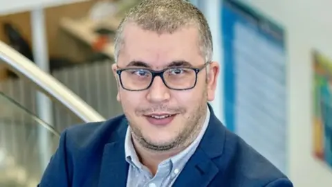 University of Southampton Professional head-and-shoulders portrait of Dr Salah Elias wearing dark-framed glasses, a blue suit jacket, and a light-colored button-down shirt. He is standing indoors, facing the camera. The background is softly blurred and includes a staircase railing and office-style interior elements in neutral and blue tones.